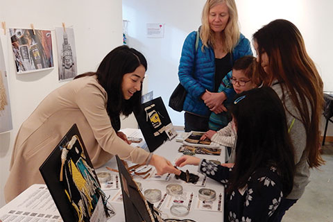 A person volunteering at an ArtStarts event stands at a table and points to items on it in front of a group of young learners