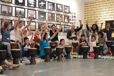 A group of young people and adults in the ArtStarts Gallery gathered around drums with their arms in the air