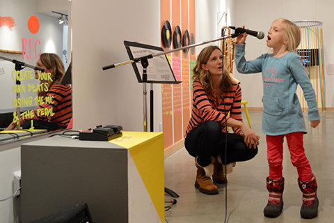 A young person sings into a microphone while an adult is crouched next to them in the ArtStarts Gallery