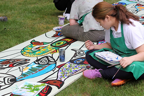 A couple young people sit on the ground and paint a drawing of several fish